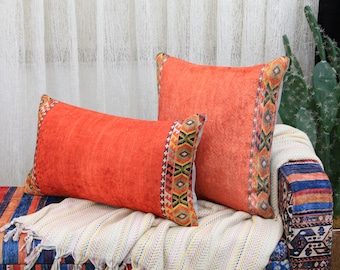 May include: Two orange throw pillows with a colorful geometric pattern border. The pillows are on a striped couch with a white throw blanket.