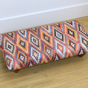 Puede incluir: Un reposapiés colorido con un patrón geométrico en tonos naranja, azul y marrón. El reposapiés tiene una forma rectangular y patas de madera.