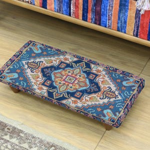 Może przedstawiać: Prostokątny podnóżek z bogatym, kolorowym wzorem w odcieniach niebieskiego, pomarańczowego i beżowego. Podnóżek wsparty jest na czterech krótkich, drewnianych nóżkach. Projekt ma estetykę orientalnego dywanu.