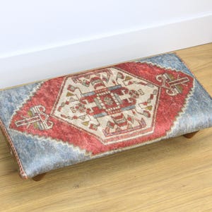 May include: Rectangular footstool with a traditional rug design. The central pattern is a red and cream geometric design, framed by a light blue border. The stool rests on four small wooden legs.