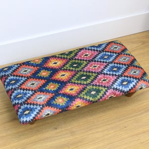 May include: A colorful patterned ottoman with a blue, red, green, and yellow geometric design. The ottoman has a wooden frame and legs.