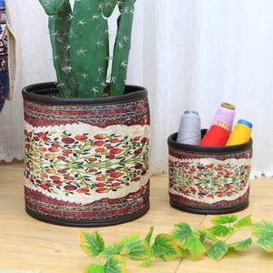 Puede incluir: Dos cestas de almacenamiento cilíndricas con un patrón floral. La cesta más grande es marrón con un fondo crema y un diseño floral rojo, verde y marrón. La cesta más pequeña es marrón con un fondo crema y un diseño floral rojo, verde y marrón. Ambas cestas tienen un ribete de cuero marrón.