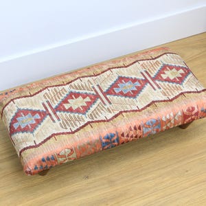Puede incluir: Un reposapiés rectangular con una cubierta de tela estampada de colores. La tela presenta un diseño geométrico con colores rojo, azul y beige.