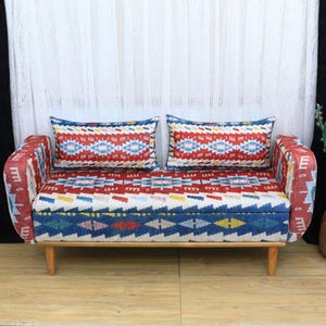 Peut inclure: Un petit canapé deux places avec un motif géométrique coloré dans les tons de rouge, bleu et blanc. Le canapé a des pieds en bois et deux coussins assortis avec le même motif.