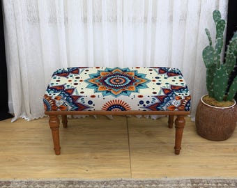 Può includere: Un pouf rettangolare con struttura in legno e quattro gambe tornite. La parte superiore imbottita presenta un colorato design a mandala nei toni dell'arancione, del blu e del bianco. Il pouf è un elemento d'arredo decorativo.