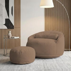 Pode incluir: Uma poltrona puff de veludo cotel&ecirc; marrom com um pufe a condizer. A cadeira tem uma forma arredondada com uma almofada. O pufe &eacute; circular. Uma moderna lumin&aacute;ria de ch&atilde;o e uma mesa lateral tamb&eacute;m est&atilde;o na sala.