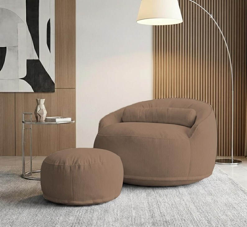 Pode incluir: Uma poltrona puff de veludo cotel&ecirc; marrom com um pufe a condizer. A cadeira tem uma forma arredondada com uma almofada. O pufe &eacute; circular. Uma moderna lumin&aacute;ria de ch&atilde;o e uma mesa lateral tamb&eacute;m est&atilde;o na sala.
