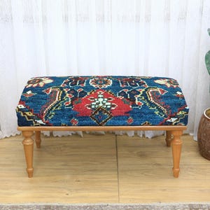 May include: A rectangular ottoman with a patterned blue, red, and cream fabric top. The ottoman has four turned wooden legs. A cactus in a brown pot is to the right of the ottoman.