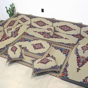 Puede incluir: Juego de asientos de suelo beige con intrincados patrones rojos, azules y dorados. El conjunto incluye múltiples cojines, una alfombra grande y algunas almohadas decorativas más pequeñas. El diseño es simétrico y presenta motivos florales y geométricos.