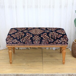 May include: A rectangular ottoman with a dark blue patterned top and light brown wooden legs. The floral pattern includes shades of brown, red, and cream. The ottoman is a decorative furniture piece.