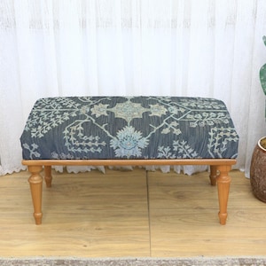 May include: A rectangular upholstered ottoman with a detailed floral pattern in shades of blue, green, and cream. The ottoman sits on four turned wooden legs. The background includes a white curtain and a potted cactus.