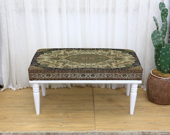 Può includere: Un pouf rettangolare con struttura in legno bianco e quattro gambe corte e arrotondate. La parte superiore è rivestita con un tessuto fantasia nei toni del beige, marrone e verde. A destra, un cactus decorativo in un vaso marrone.