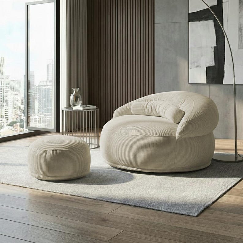 Pu&ograve; includere: Una poltrona in velluto a coste beige con un pouf abbinato. La poltrona arrotondata ha schienale e bracciolo curvi e un cuscino cilindrico. Il pouf ha una forma bassa e circolare. L'arredamento &egrave; su un tappeto grigio chiaro.