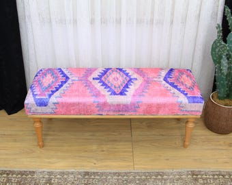 May include: A rectangular bench with a pink, purple, and blue geometric patterned top. The bench has light brown wooden legs and a matching wooden frame. The bench is a decorative piece of furniture.