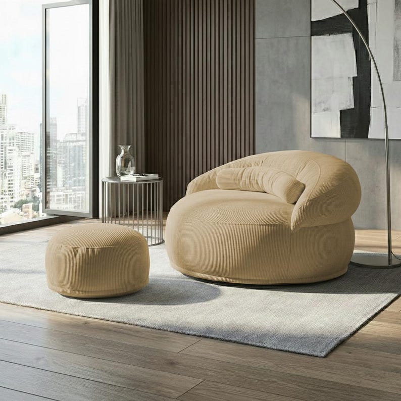 May include: A beige corduroy armchair with a matching ottoman. The rounded armchair has a curved back and armrest, and a cylindrical bolster pillow. The ottoman is a low, circular shape. The furniture sits on a light gray rug.