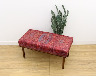 May include: A red and blue patterned bench with wooden legs. The bench is sitting on a wooden floor.
