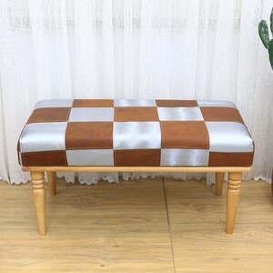 Puede incluir: Un banco tapizado con estampado de cuadros marrones y plateados con patas de madera. El banco está sobre un suelo de madera.