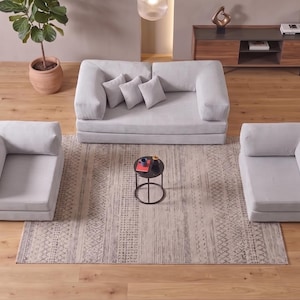 Puede incluir: Un conjunto de sofá modular gris claro que incluye un sofá ancho de dos plazas con tres cojines a juego y dos sillones sin brazos. Una pequeña mesa auxiliar redonda de metal negro con una superficie oscura se sitúa en el centro.