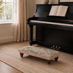 May include: A rectangular piano footstool featuring a beige, cream, and dark blue patterned fabric top with a central medallion design. The footstool is supported by four short, dark brown, rounded wooden legs, positioned on a floor.