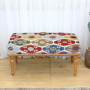May include: A rectangular ottoman with a patterned fabric top in red, blue, yellow, and brown geometric designs. The wooden legs are light brown. The ottoman is a decorative furniture piece.
