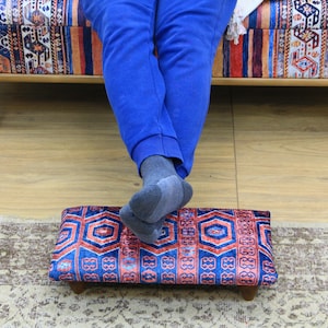 May include: A blue and red patterned footstool with wooden legs. The footstool is sitting on a rug.