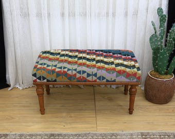 May include: A rectangular upholstered bench with a geometric pattern in shades of red, yellow, blue, and purple. The bench has four wooden legs and a dark brown frame. The bench is a decorative furniture piece.