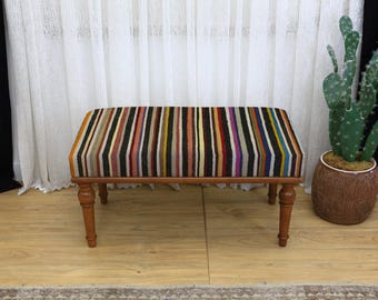 May include: A rectangular bench with a multi-colored striped top and wooden legs. The stripes include shades of yellow, orange, red, purple, and black. The bench is placed on a light-colored wooden floor.