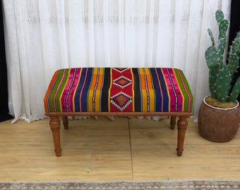 May include: A rectangular bench with a colorful, patterned fabric top. The fabric features vertical stripes in various colors, including pink, green, orange, and blue, with a central red diamond design. The bench has four wooden legs.