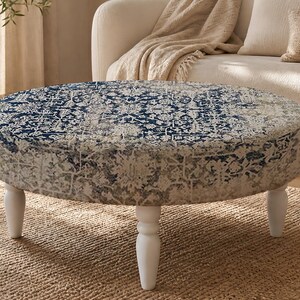 May include: A round ottoman featuring a blue and beige intricate patterned fabric upholstery. The piece is supported by four short, white, turned wooden legs that provide a stable base for the circular, cushioned top surface.