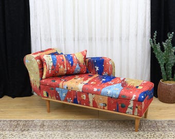 Könnte beinhalten: Eine Chaiselongue mit einem farbenfrohen abstrakten Muster in Rot, Blau und Gelb. Der Holzrahmen hat vier Beine. Ein passendes Kissen liegt auf dem Sitz. Ein Kaktus in einem braunen Topf befindet sich im Hintergrund.