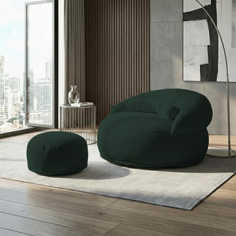 Pode incluir: Um conjunto de poltrona e pufe a condizer, verde escuro, com textura de veludo cotel&ecirc;. A poltrona arredondada tem um encosto baixo e um assento profundo. O pufe &eacute; circular e fica rente ao ch&atilde;o. O conjunto est&aacute; sobre um tapete cinzento claro.