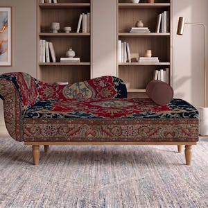 May include: A chaise lounge featuring a rolled armrest and a cylindrical bolster pillow, upholstered in a fabric with a dark red, navy blue, and beige intricate floral pattern, supported by four light-colored wooden legs with a turned design.