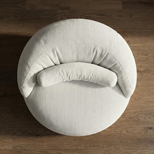 May include: Overhead view of a round, off-white corduroy chair with a curved backrest and a cylindrical bolster pillow. The chair is set on a dark wood floor, creating a contrast with the light-colored fabric.