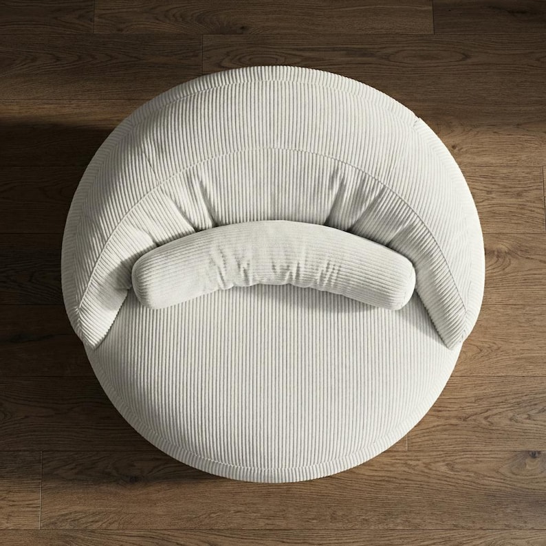 May include: Overhead view of a round, off-white corduroy chair with a curved backrest and a cylindrical bolster pillow. The chair is set on a dark wood floor, creating a contrast with the light-colored fabric.
