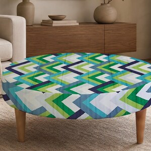 May include: A round ottoman featuring a geometric chevron pattern in shades of navy blue, bright green, light green, teal, and white. The piece is supported by four light-colored wooden legs that provide a stable base for the circular upholstered seat.