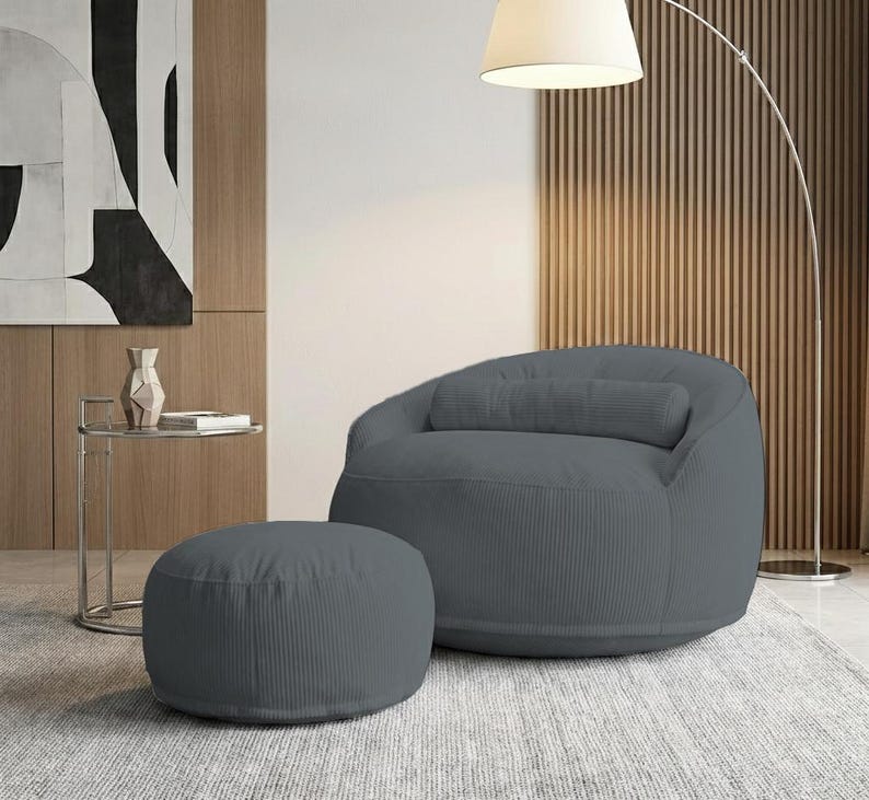 Pu&ograve; includere: Una poltrona a sacco in velluto a coste grigio con un pouf abbinato. La sedia ha una forma arrotondata con un poggiatesta integrato. Il pouf &egrave; circolare. L'arredamento &egrave; in un ambiente moderno.