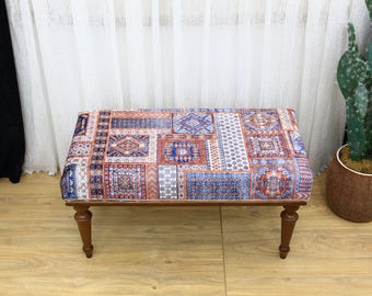May include: A wooden bench with a patterned fabric seat. The fabric is a patchwork of different colors and designs, including blue, red, and white. The bench has four legs and is sitting on a wooden floor.