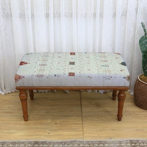 May include: A rectangular ottoman with a patterned fabric top and wooden legs. The fabric features a light background with a repeating design in red, blue, and brown. The legs are a warm brown colour.