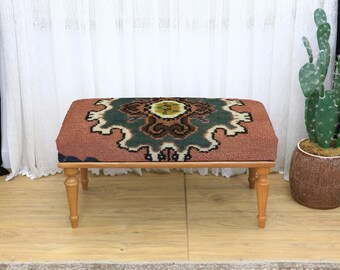 May include: A rectangular ottoman with a patterned top in shades of rust, green, and cream. The ottoman has a wooden frame and four short, turned legs. The design on the top is symmetrical and intricate, adding a decorative element to the furniture.
