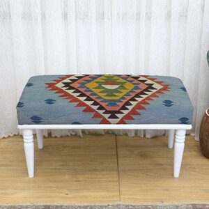 May include: A rectangular bench with a white wooden frame and four legs. The seat is upholstered with a blue fabric featuring a geometric pattern in red, yellow, and brown. A cactus in a brown pot is to the right.
