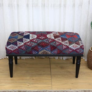 May include: A patterned fabric ottoman in red, blue, and white, with a black wooden frame and four turned legs. The ottoman is a decorative furniture piece, suitable for a living room or bedroom.
