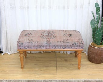May include: A rectangular ottoman with a patterned, faded pink and green fabric top. The ottoman has four turned wooden legs. A cactus in a brown pot is to the right of the ottoman.