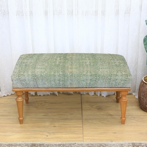 May include: A rectangular upholstered bench with a light green patterned fabric top and four wooden legs. The bench is set on a light wood floor, with a cactus in a brown pot to the right.