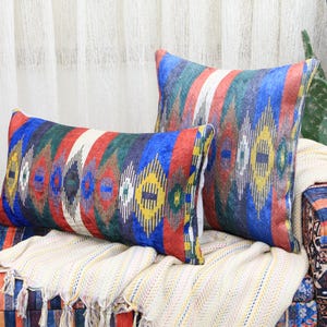 Puede incluir: Dos almohadas decorativas de colores con diseños geométricos en tonos azules, rojos, amarillos y verdes. Las almohadas están sobre un sofá a rayas con una manta blanca.