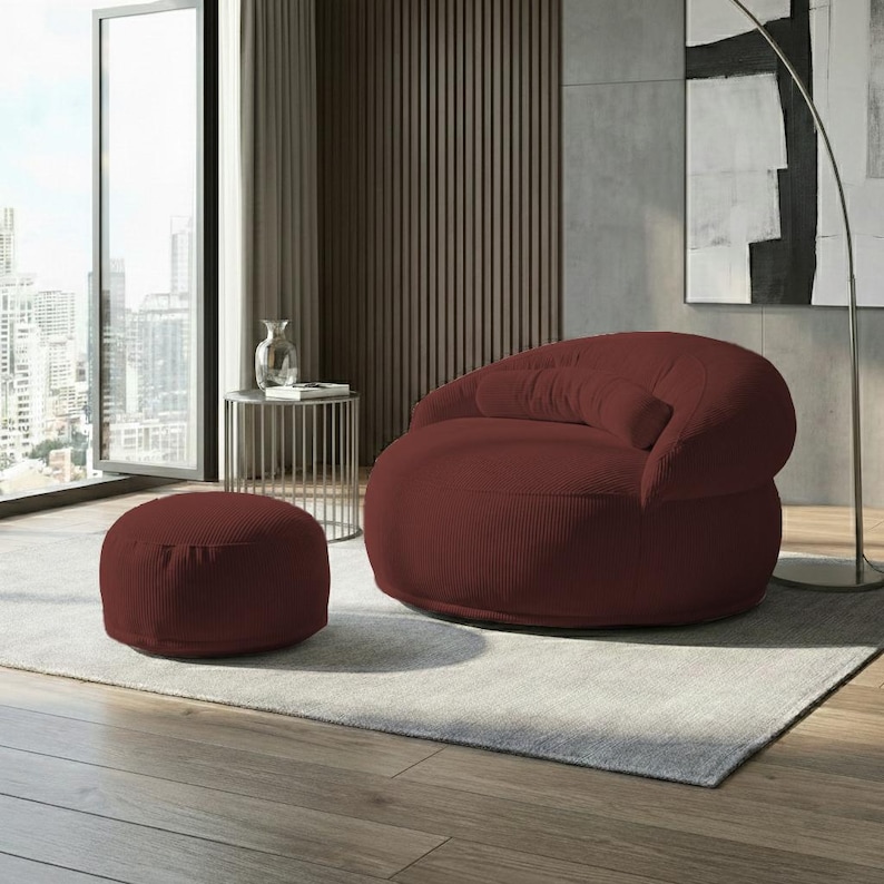 May include: A burgundy corduroy armchair and matching ottoman set. The rounded armchair has a deep seat and a curved back. The ottoman is circular and low to the ground. The set sits on a light gray rug in a modern living space.