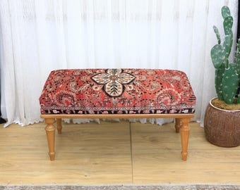 Puede incluir: Un otomán rectangular con una parte superior estampada con flores rojas, negras y crema. El otomán tiene cuatro patas de madera de color claro. El diseño de la parte superior recuerda a una alfombra tradicional. Una pieza decorativa para una sala de estar o un dormitorio.