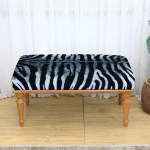 May include: A rectangular bench with a zebra print cushion. The cushion features black and white stripes. The bench has four wooden legs and a light brown frame. A cactus in a brown pot is to the right.