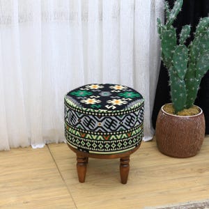 Può includere: Un pouf rotondo nero e verde con motivo geometrico e gambe in legno. Il pouf presenta un design geometrico con accenti neri, verdi, bianchi e arancioni.