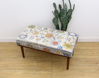 Puede incluir: Un banco de madera con un cojín de estampado floral. El cojín tiene un fondo blanco con flores azules, rojas y amarillas. El banco tiene un marco de madera marrón oscuro.
