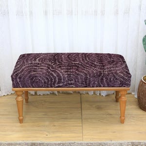 May include: A rectangular upholstered bench with a patterned, dark purple top and light brown wooden legs. The bench is a decorative furniture piece, suitable for a living room or bedroom. The legs are slightly curved.